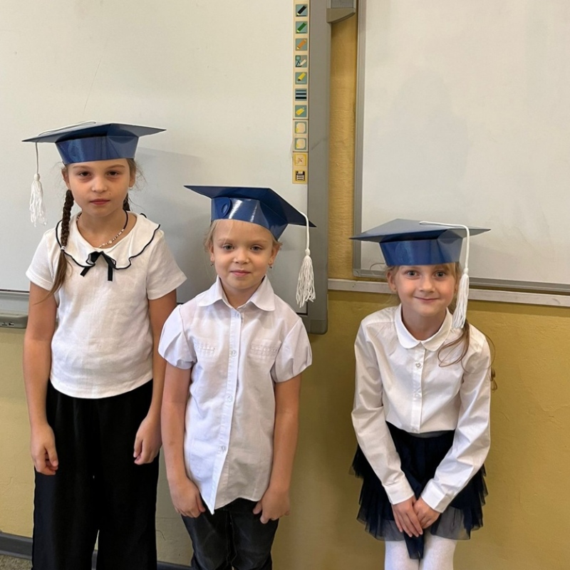 3 dziewczynki stoją w klasie przed białą tablicą, uśmiechnięte i w biretach