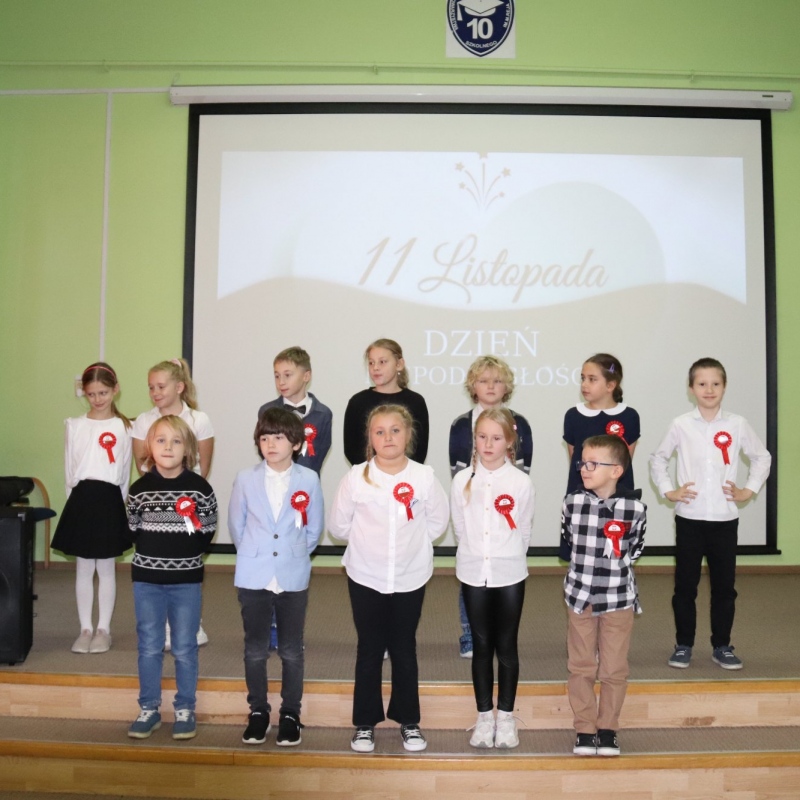 Uczniowie klasy 2a na scenie z kotylionami na wyswietlanym tle z flaga i napisem 11 listopada