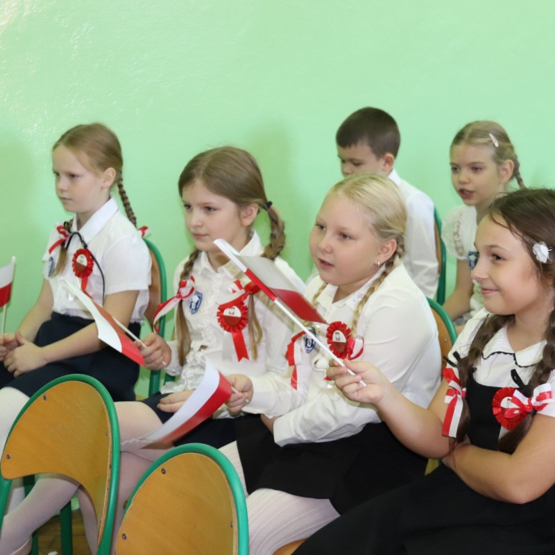 Uczniowie siedzą mają w rękach polskie flagi, ubrani na galowo