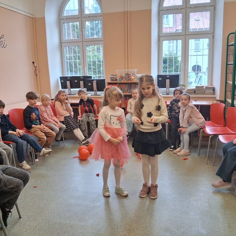 uczniowie siedzą w kole w bibliotece, dwie uczennice stoją na środku